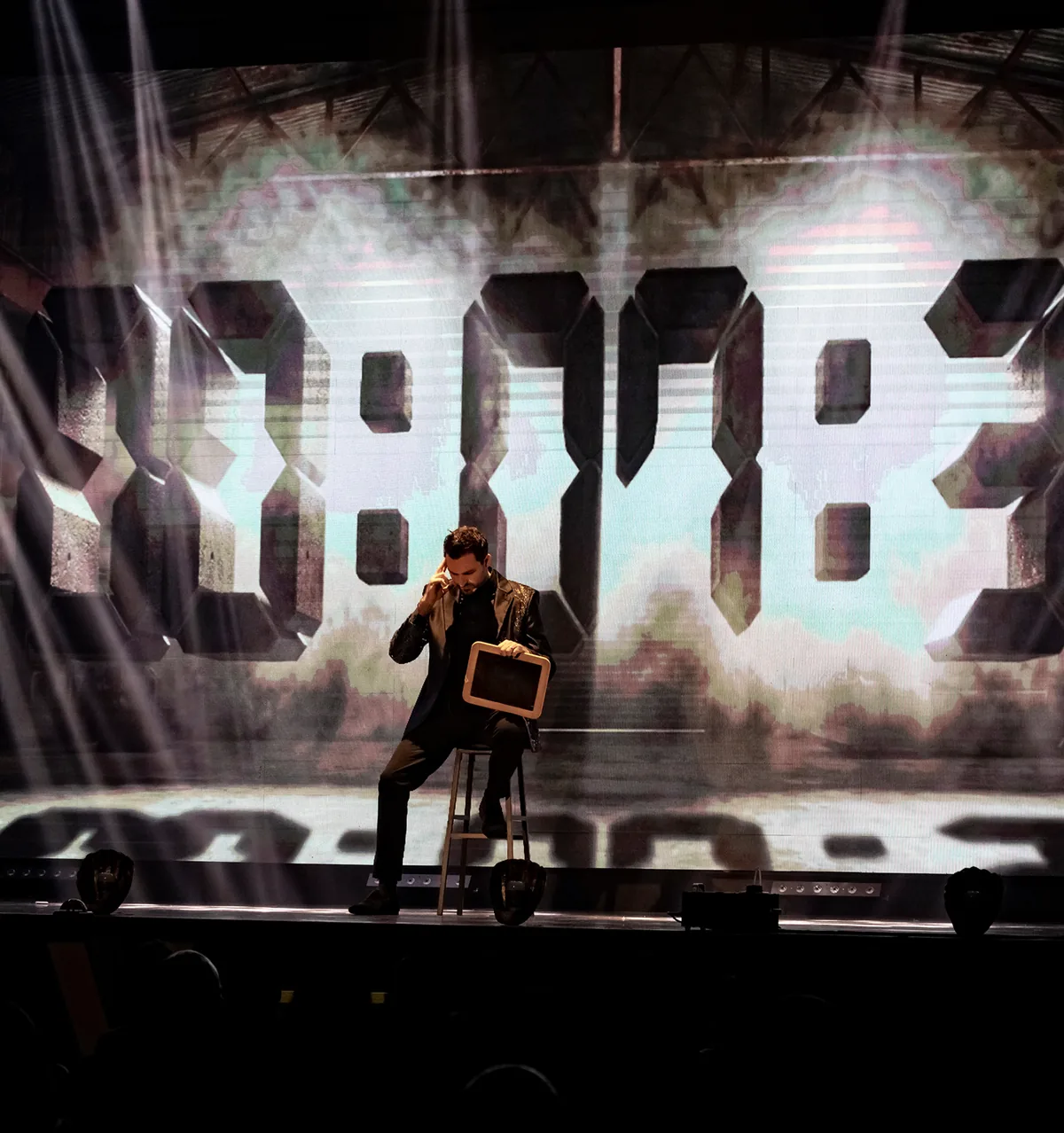 Fernando Mier seated on a stool on stage, holding a slate with a large screen behind him.