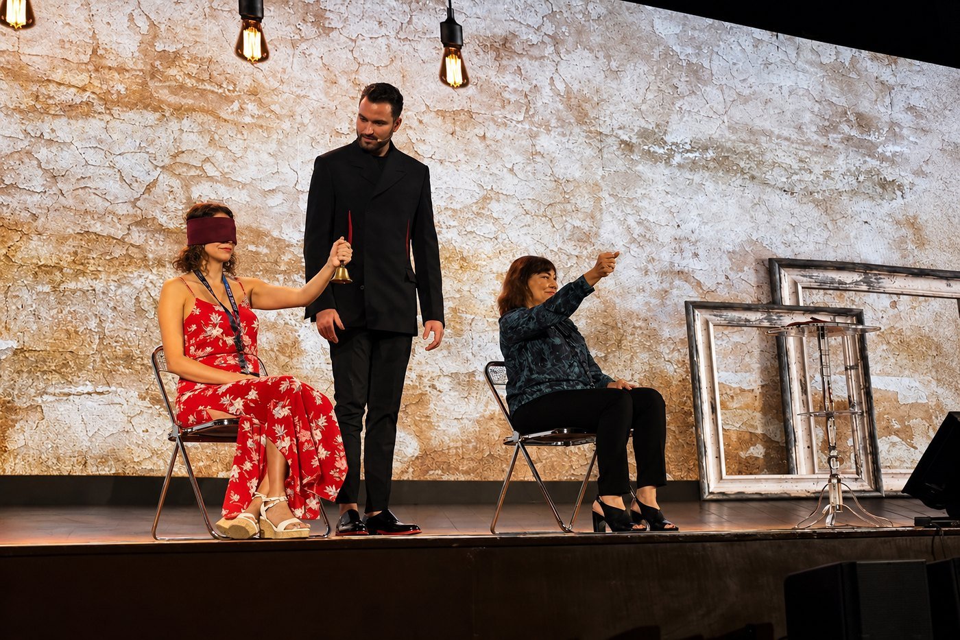 Fernando Mier junto a dos voluntarias en el escenario; una de ellas, con los ojos vendados, sostiene una campana